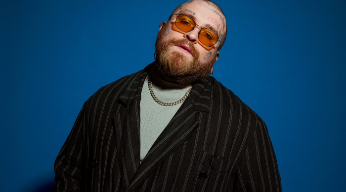 man with face tattoos, a ginger beard and sunglasses, standing against a blue background